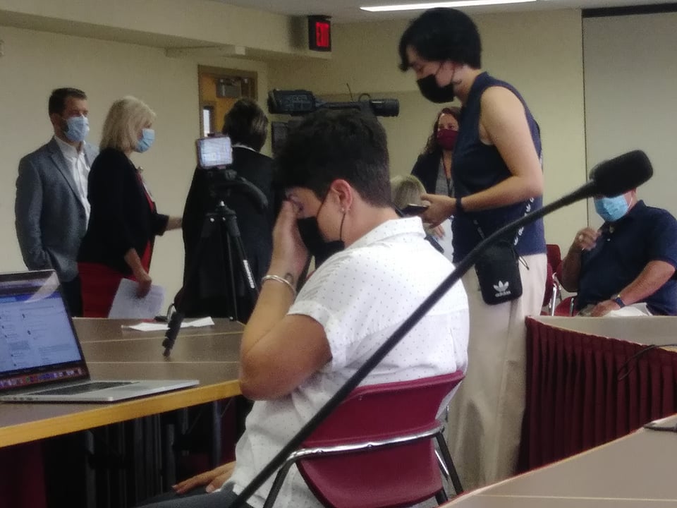 A photo from inside the Macon County Board Room, sitting in the back row. Pictured is a woman of the press rubbing her eyes or forehead, and several officials in the background appearing to converse. I'm sorry this alt text is not more descriptive, but it's not really an important photo.