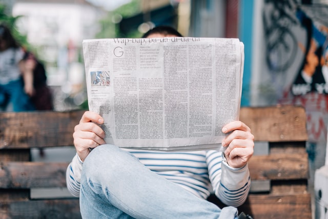 A photo of a white woman sitting on a bench, with her right ankle atop her left knee. Her arms are forward, holding a newspaper at a healthy viewing distance. She\'s wearing lightly colored denim jeans and a long sleeve shirt with blue and white horizontal stripes.